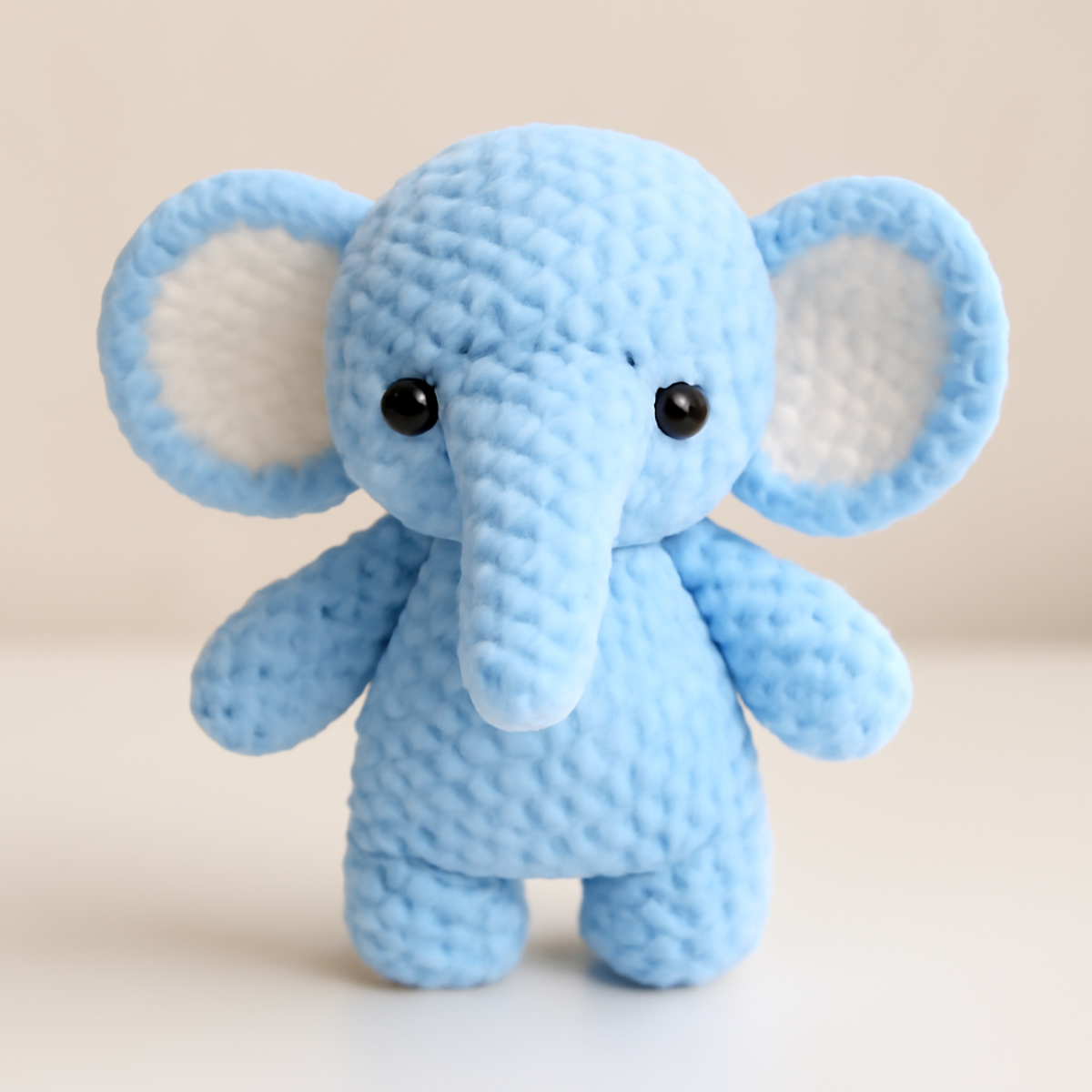 A light blue baby elephant toy made from a crochet pattern, featuring a rounded body, a long trunk, large ears with contrasting ear lining, and black safety eyes.