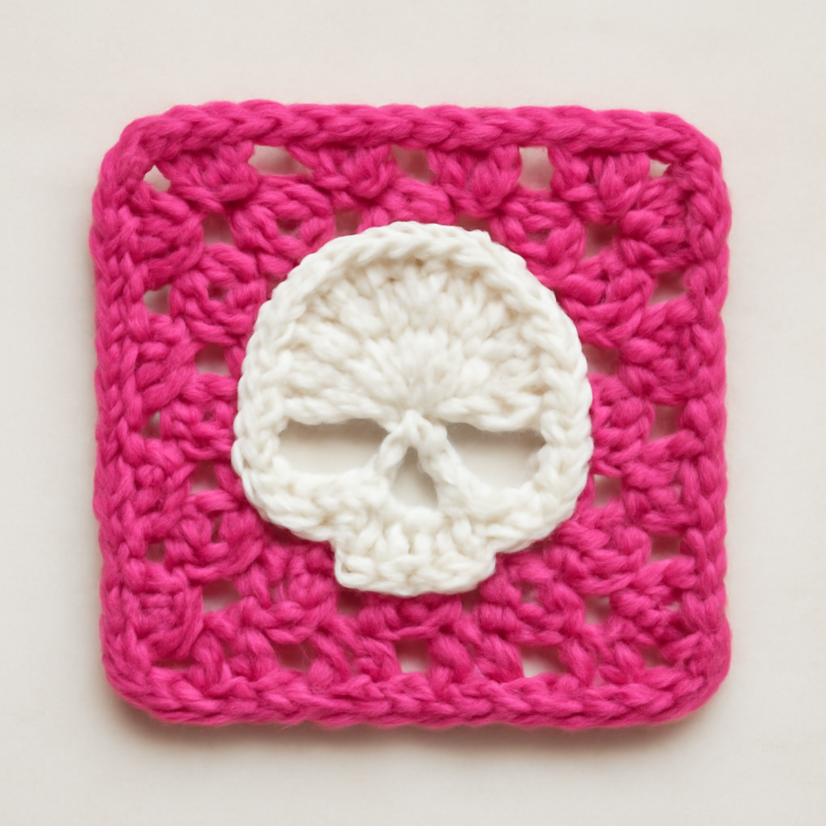 A pink and white granny square with a skull motif at the center, showcasing a slightly raised texture. The square is approximately 6 inches in size.