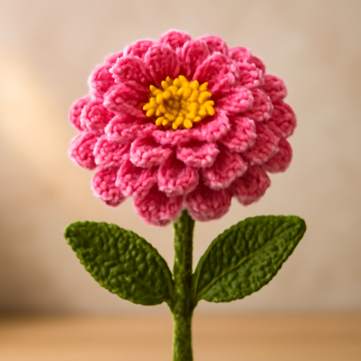 Crochet Zinnia Flower Pattern featuring a multi-layered flower in shades of pink and a yellow center, with green leaves.